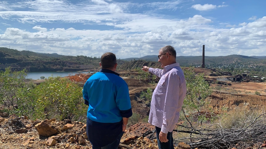 Two men, one in a blue shirt and one in a white and purple shirt, over look a mine site.