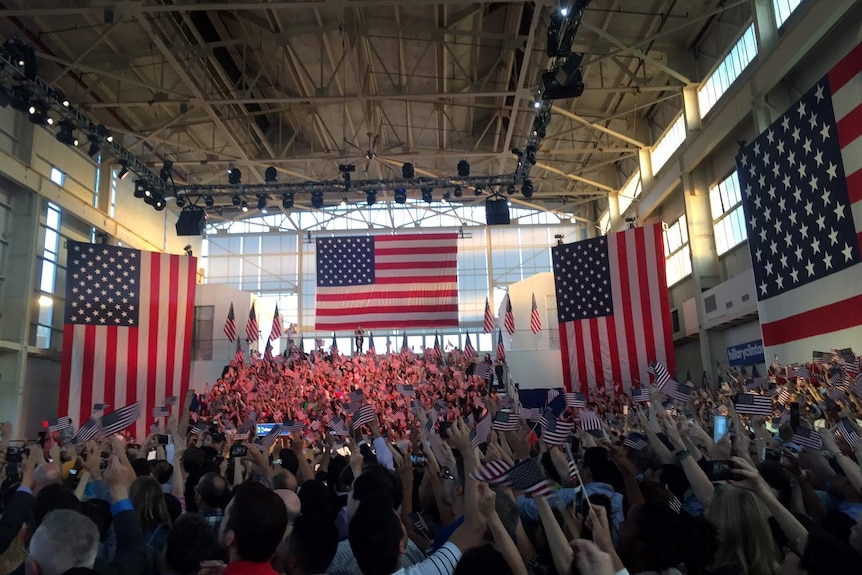 Giant American flags hang from walls of an aircraft hanger full of people also waving flags.