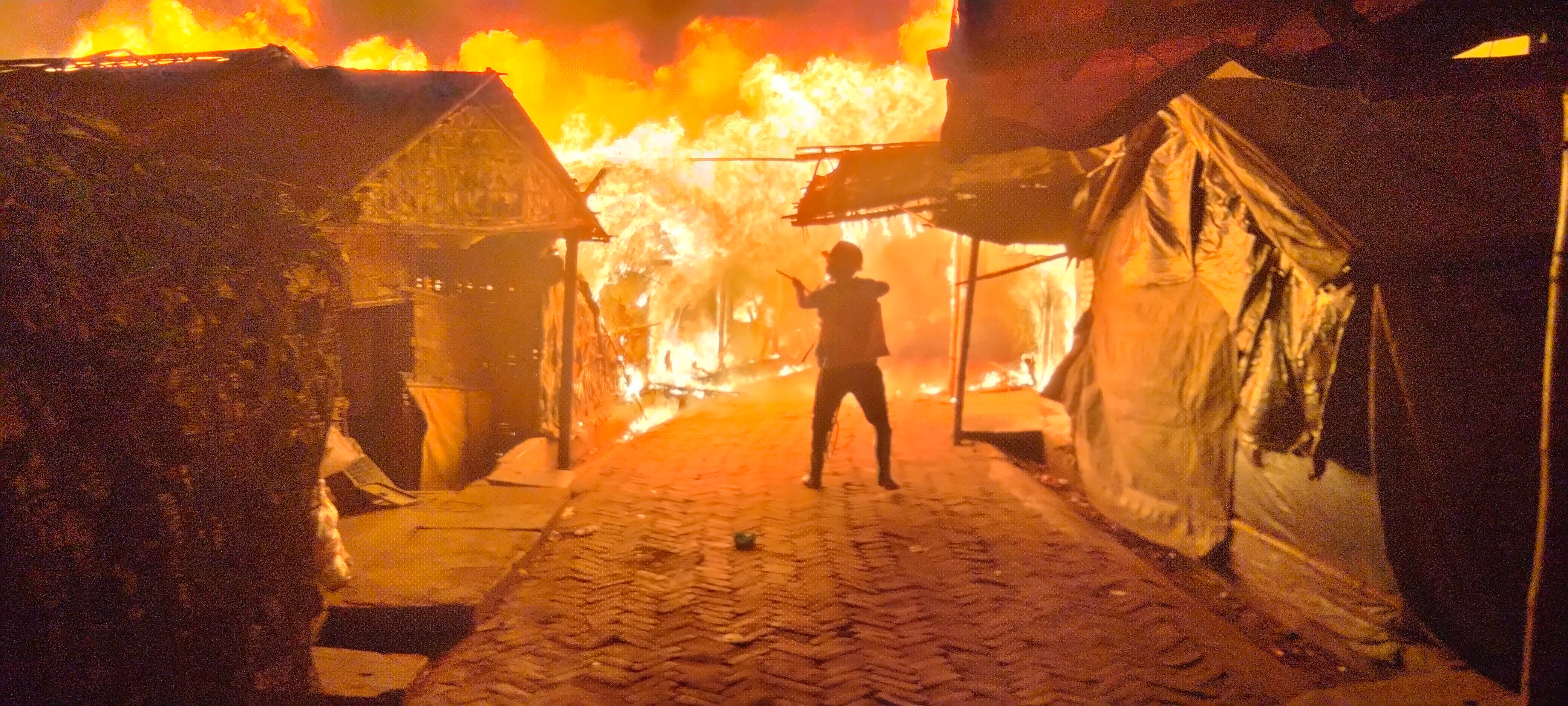 A single firefighter fights a huge fire as it destroys wooden and plastic shelters