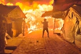 A single firefighter fights a huge fire as it destroys wooden and plastic shelters