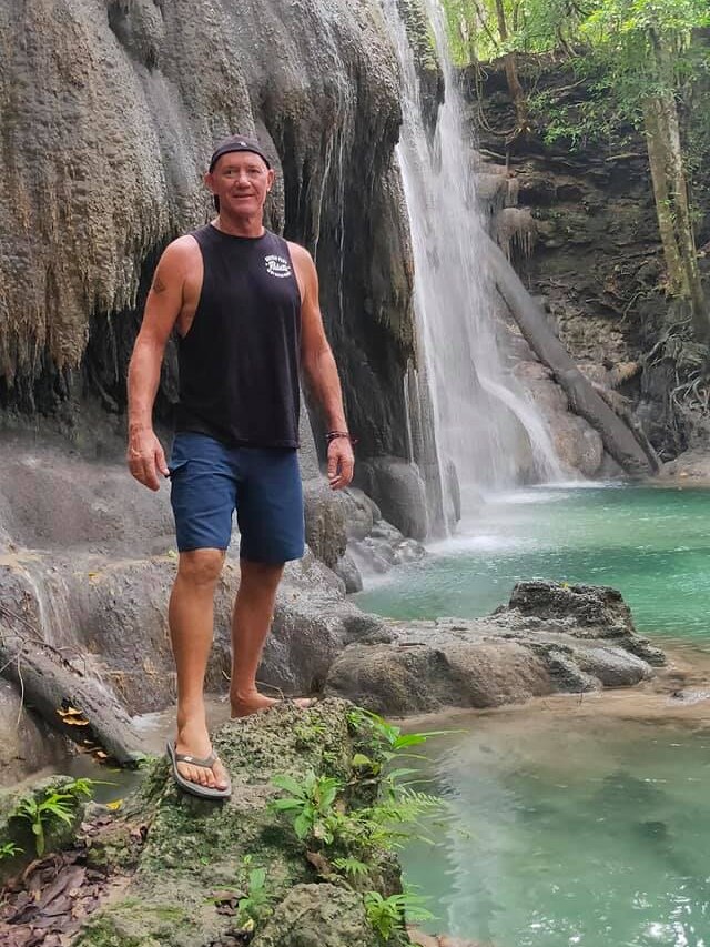 A man in a black muscle shirt and cap stands next to a waterfall 