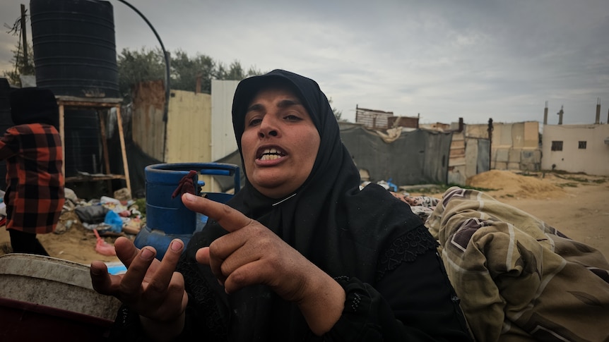 A woman wearing black hijab speaks from a cart, in the middle of an area with makeshift structures