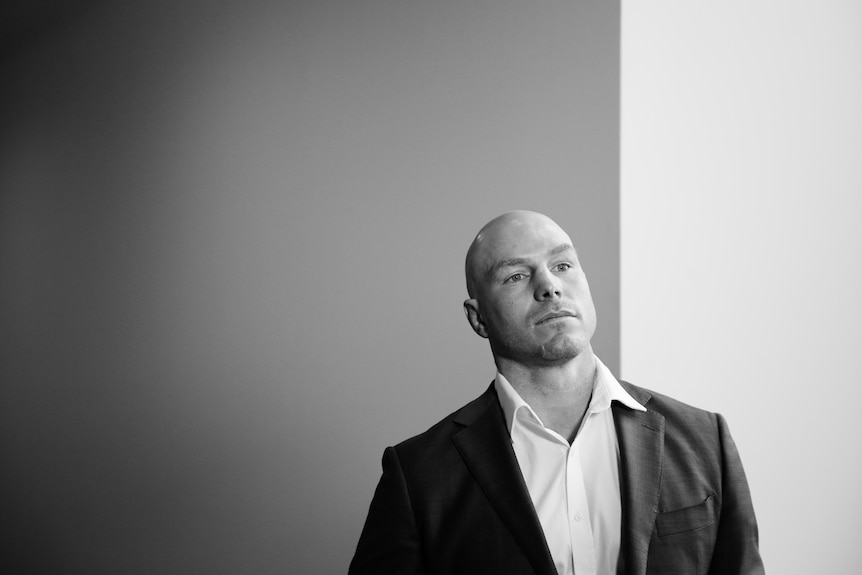 A black and white portrait of David Pocock wearing a suit standing in front of a white wall