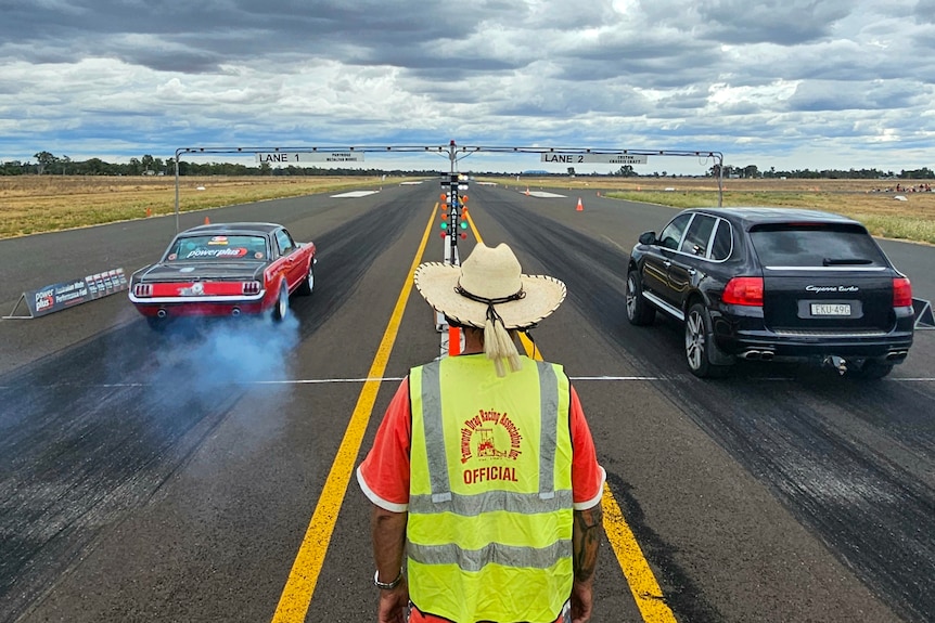 Two cars drag race down a runway, being watched by a man in an offical's vest