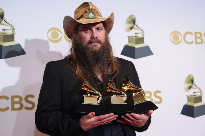 chris stapleton holds up three grammy awards standing in front of a promo banner