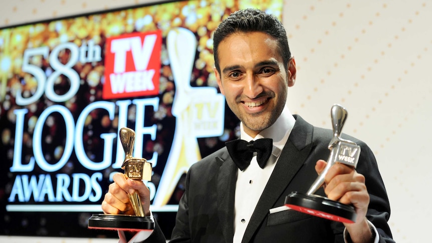 Waleed Aly holds his gold and silver Logies with a Logies sign behind him.