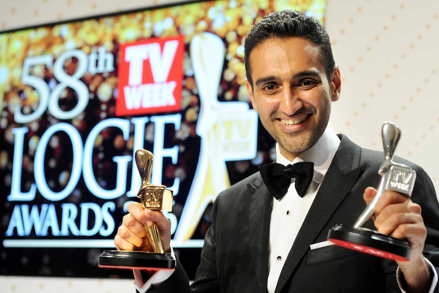 Waleed Aly holds his gold and silver Logies with a Logies sign behind him.