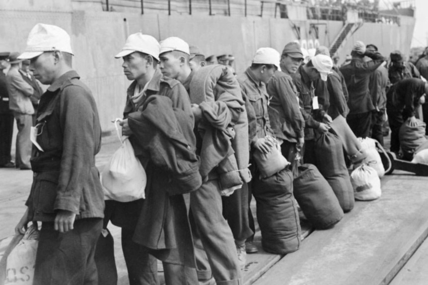 A line of Japanese prisoners of war line up holding luggage preparing to board a ship.