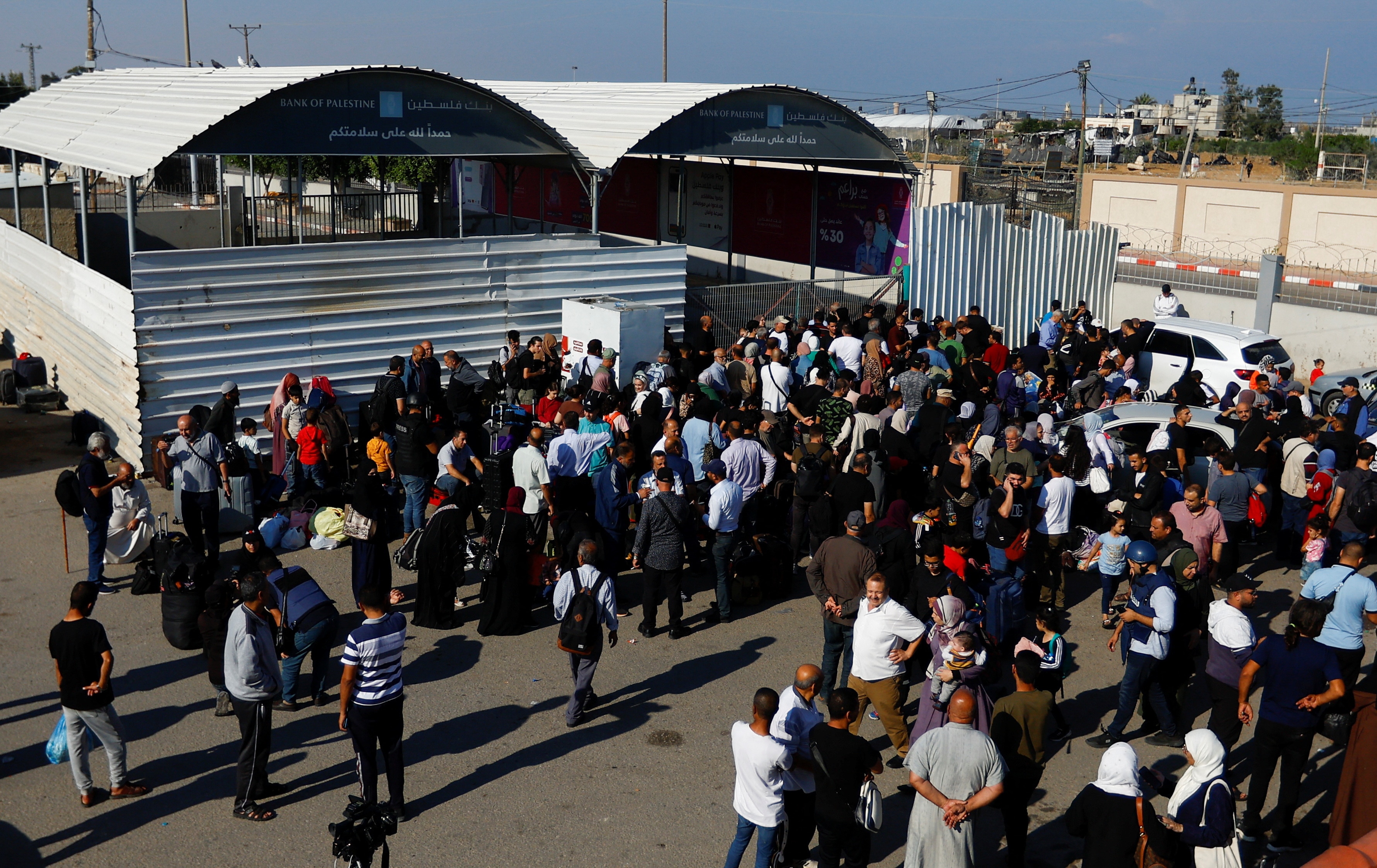 A group of Middle Eastern people stand in a car park next to a gated tin shed on sunny day