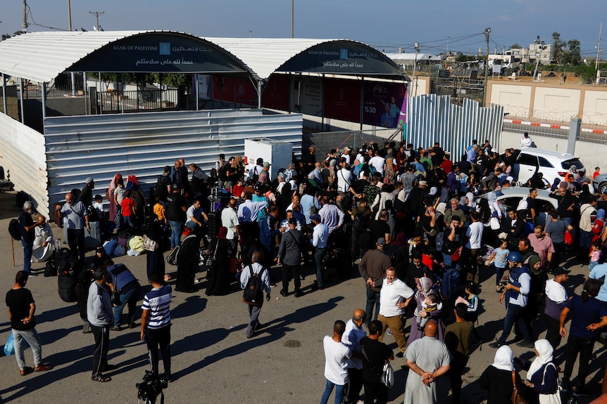 A group of Middle Eastern people stand in a car park next to a gated tin shed on sunny day