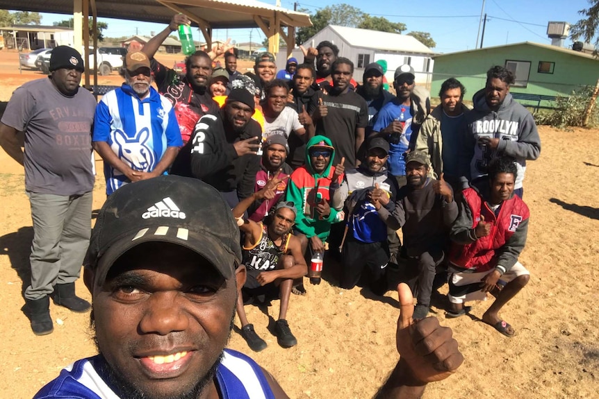 About 25 Aboriginal men, some in footy jumpers, gather for a group selfie, giving the thumbs up and other happy gestures.