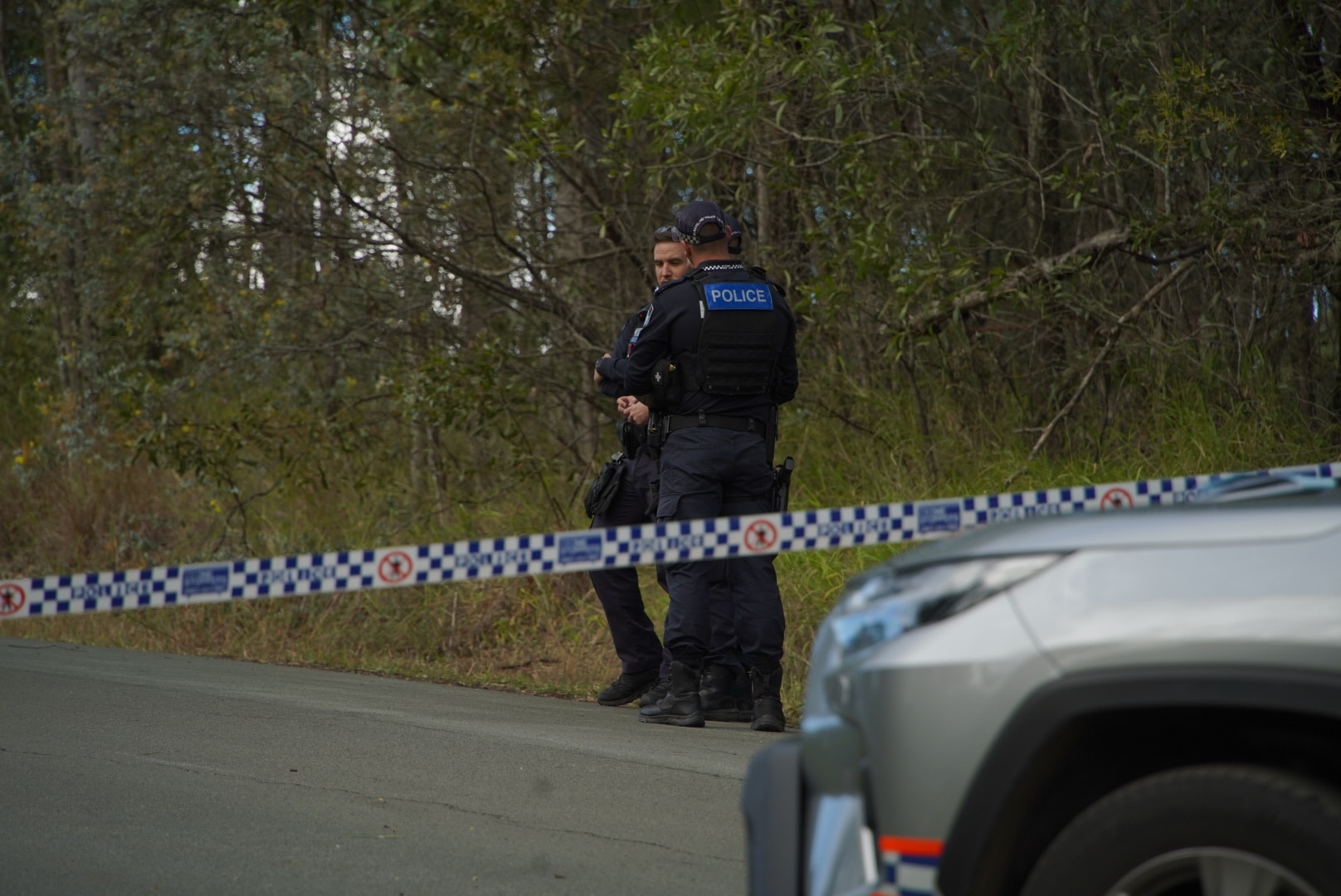 Two police officers stand at the side of a road behind police tape.