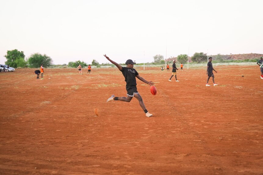 A player kicks a football at training on a red dirt oval.
