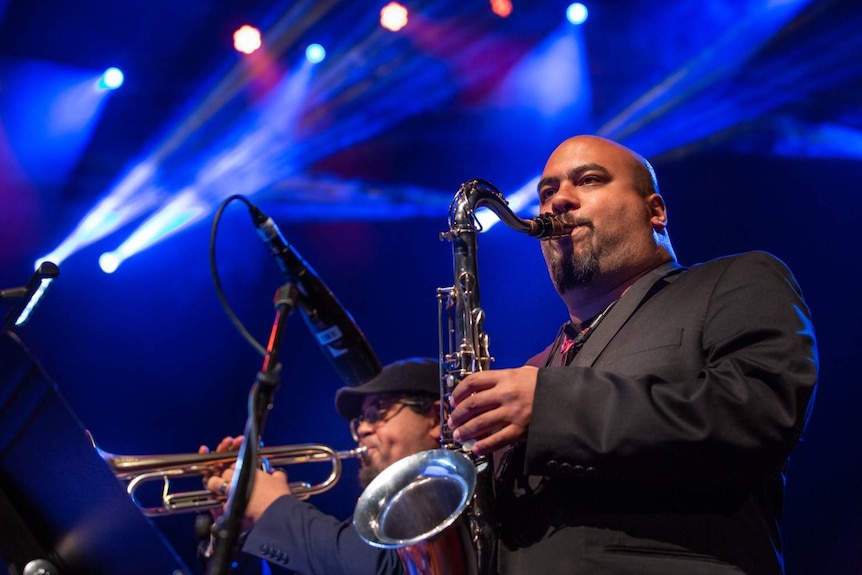 Troy plays the saxophone in front of blue lights, up on stage.