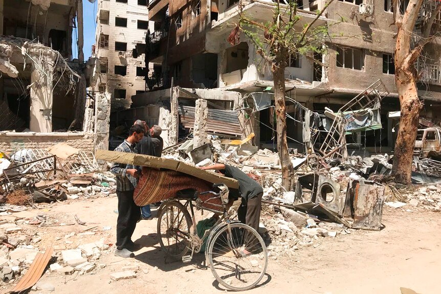 Men load a carpet and mattress on to a bicycle in front of damaged buildings in the town of Douma.