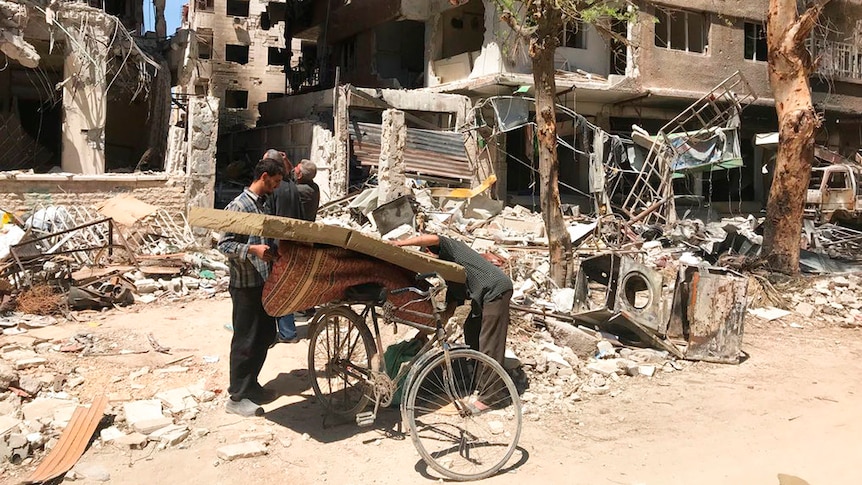 Men load a carpet and mattress on to a bicycle in front of damaged buildings in the town of Douma.