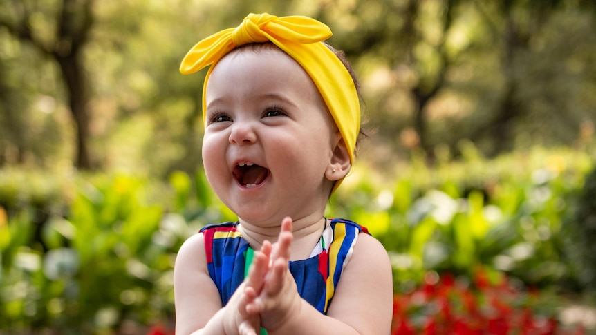 A baby girl is laughing and clapping her hands, she is wearing yellow headband and blue dress, she is sitting down.