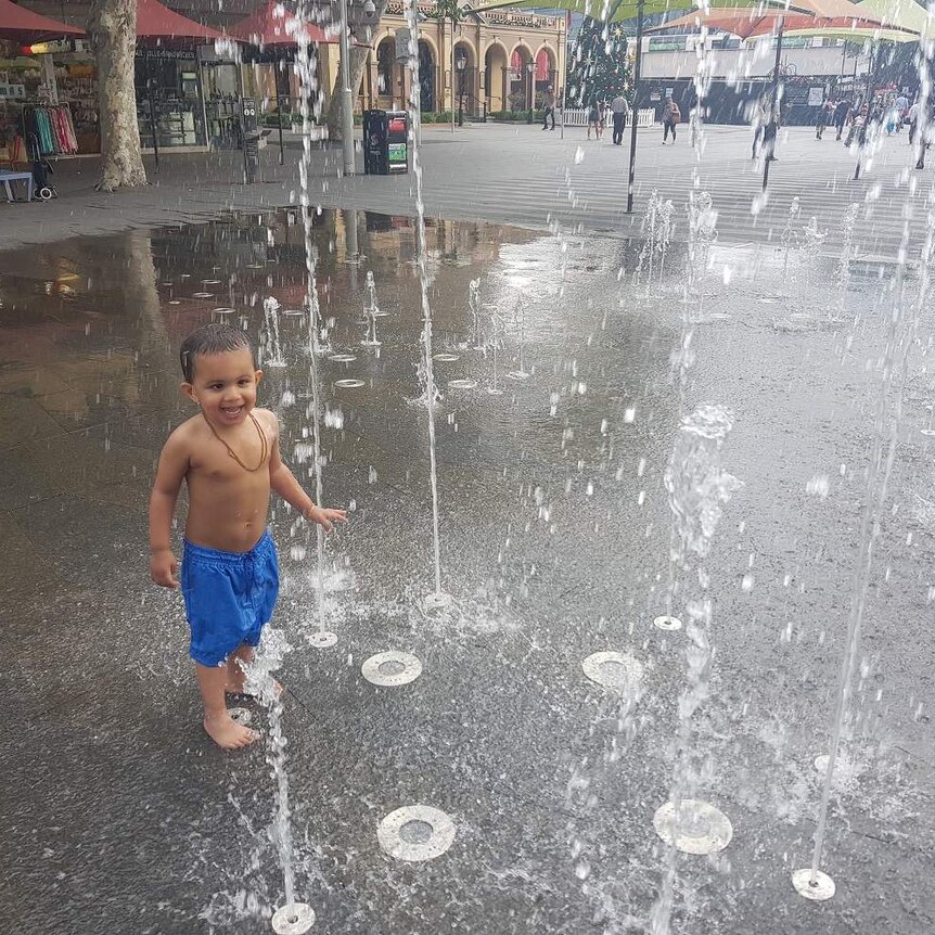 A young child in blue boardshorts cooling off under the water of a fountain in Parramatta.