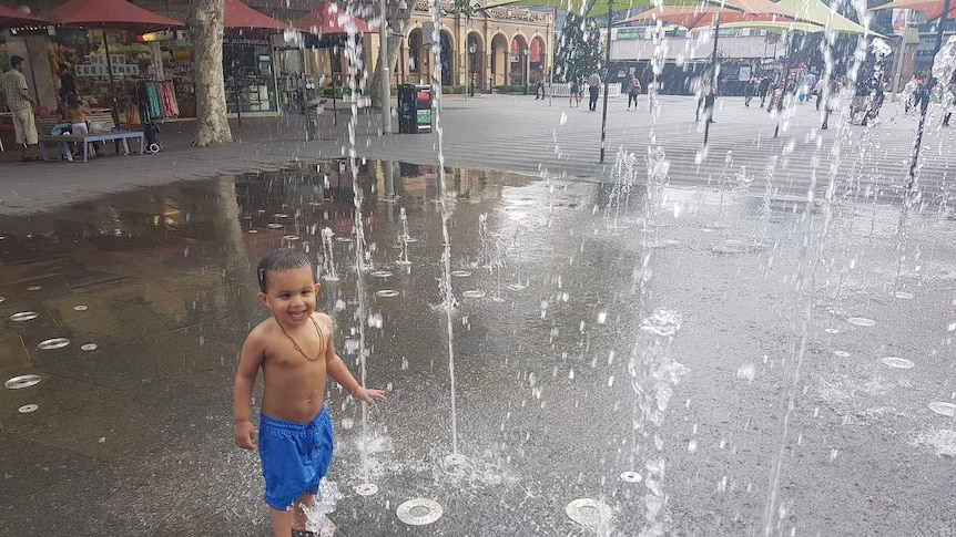 A young child in blue boardshorts cooling off under the water of a fountain in Parramatta.