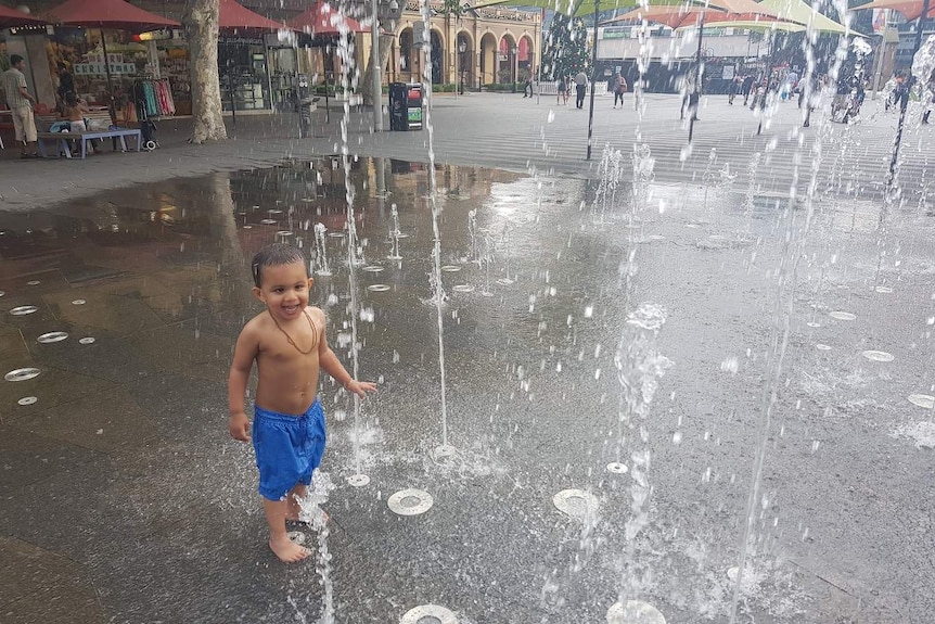 A young child in blue boardshorts cooling off under the water of a fountain in Parramatta.