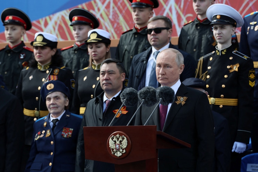 Vladmir Putin standing at a lectern, surrounded by people in formal uniforms. 