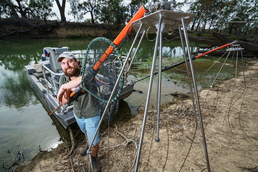 A man standing with a net next to a fishing boat in front of a river bank.