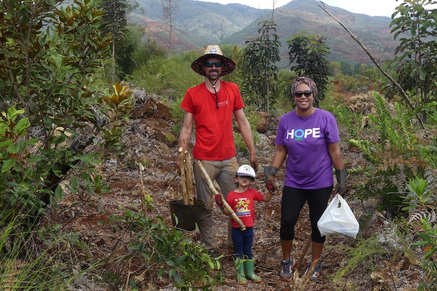 A man and woman and child with red and purple t-shirts and green trees and bush around them.
