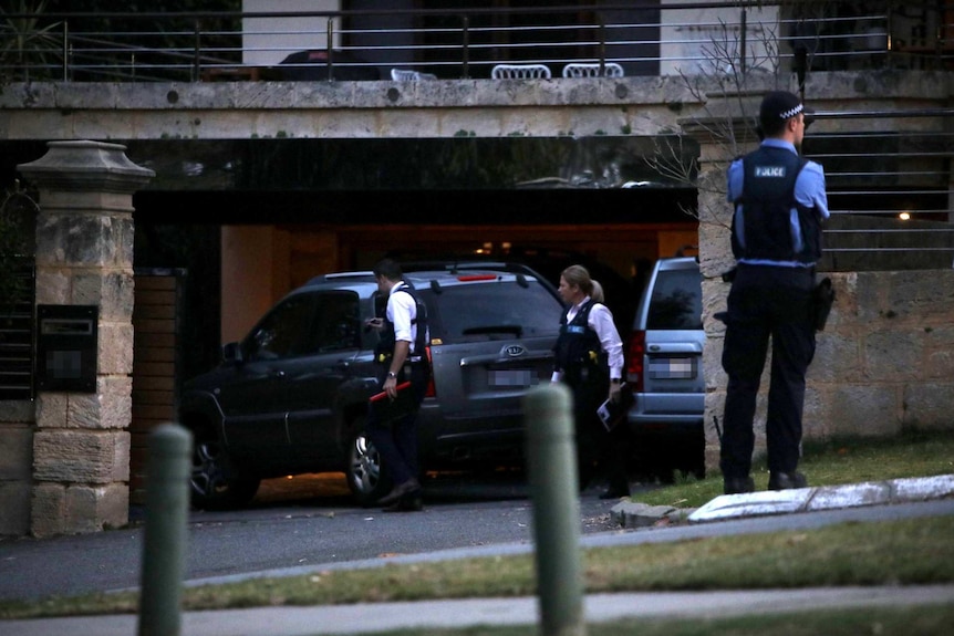 Two plain clothes police officers walk behind two cars parked in the garage of a house, with a uniformed police officer nearby.