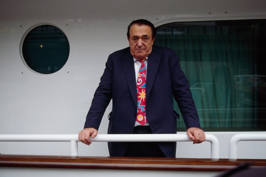 Robert Maxwell dressed in a suit with a patterned red tie leans on the railing of a boat.