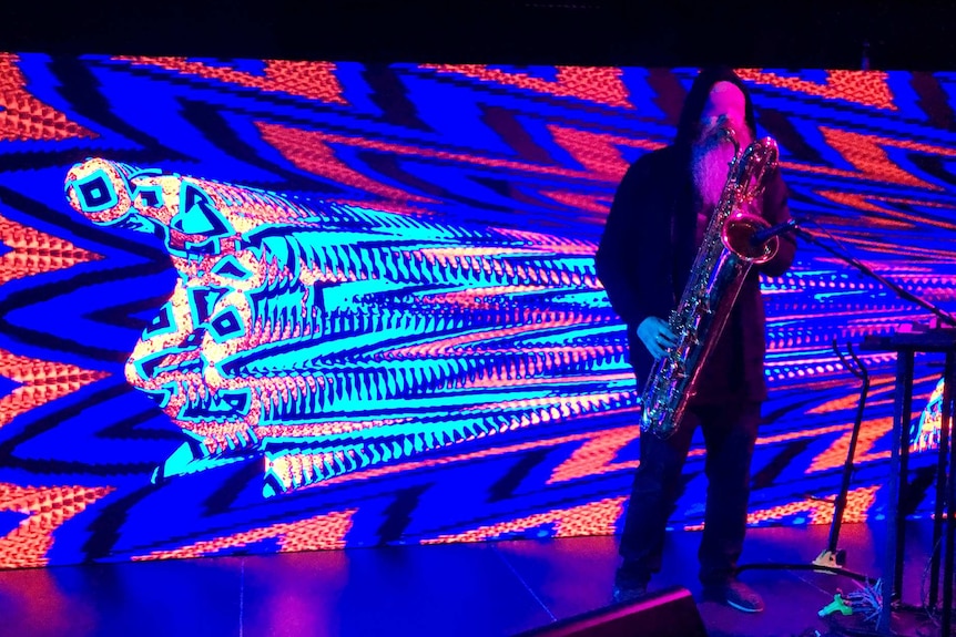 A saxophonist with taped up eyes performs in front of a colourful LED screen