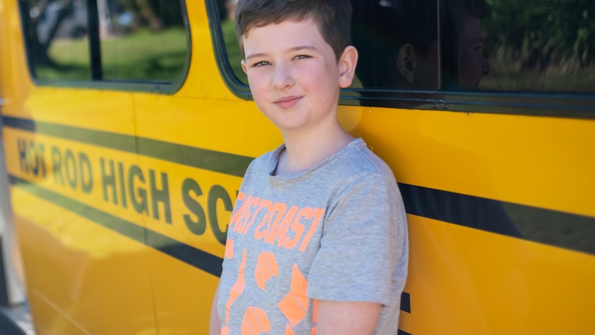 一个八岁的男孩靠在一辆亮黄色的公共汽车旁边。