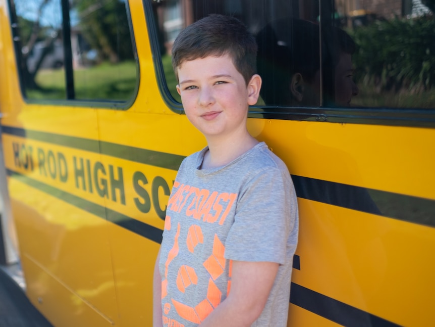 An eight year old boy leans against the side of a bright yellow bus.