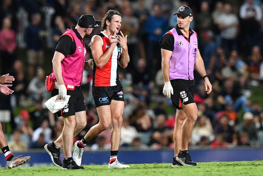 An AFL player comes off the ground holding his injured jaw as two team officials walk with him during a game.