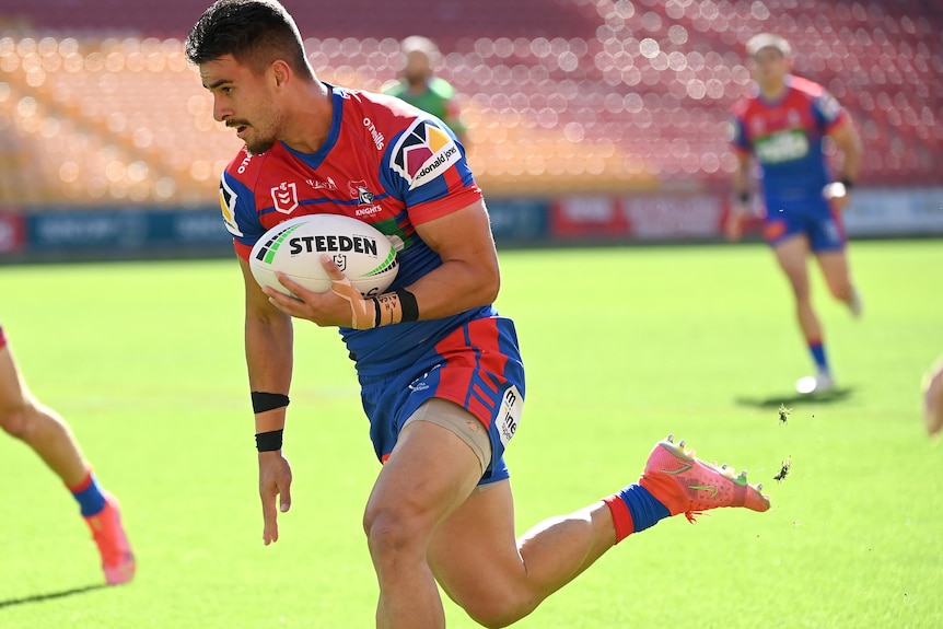 A bearded Polynesian man in red and blue jersey carries a rugby ball on grassy stadium pitch.