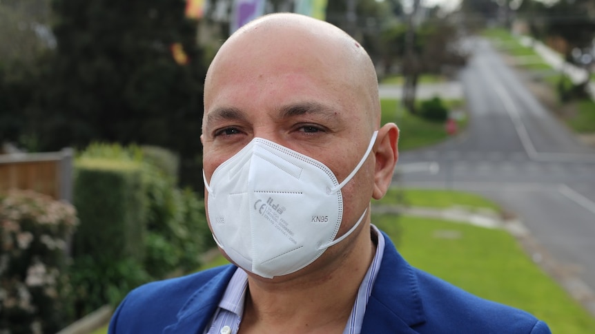 John stands on a suburban nature strip, wearing a white mask and blue suit.