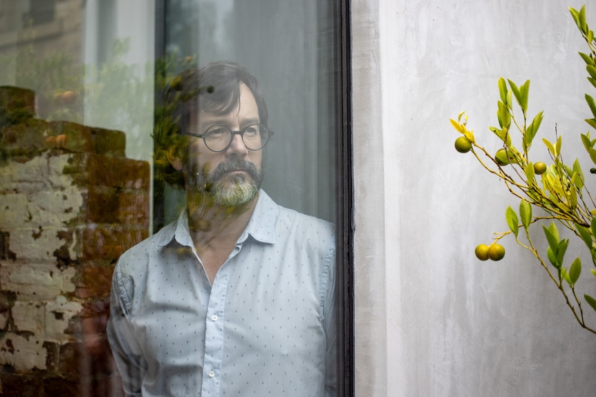 Sam stands at a large window, looking out. A fruit tree is growing in a pot outside to the right of him.