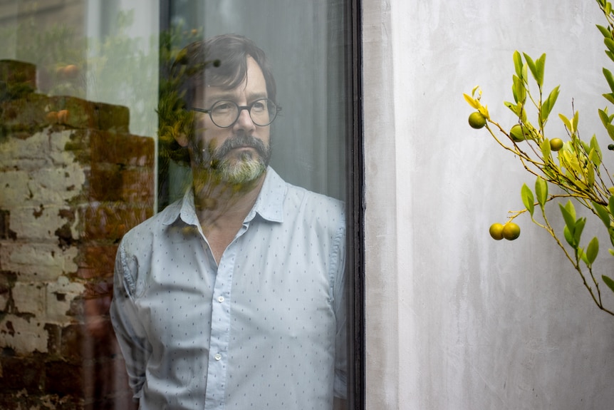 A bearded man stands behind a window