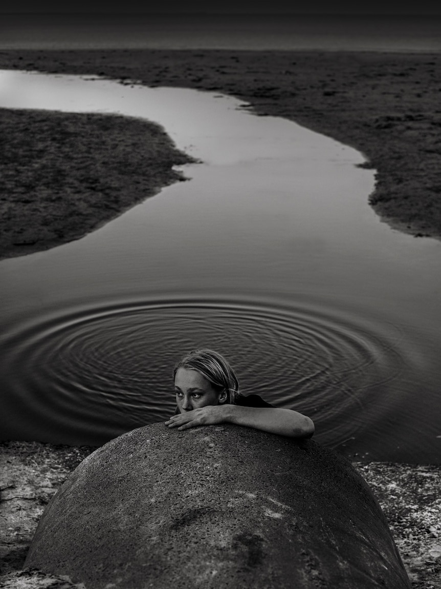 Monochrome art photograph of a boy swimming in a  storm drain, resting by holding onto the drain pipe.