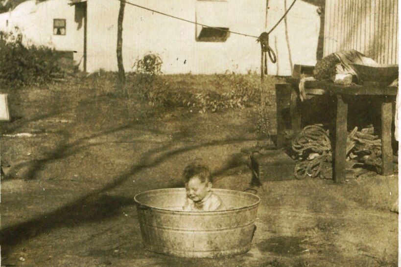 An aged sepia photo shows a small child sitting in a tin tub on the dirt. Behind them is a white tin home.