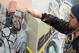 A man in a paint-splattered top uses a roller to paint over a defaced depiction of a bearded, bespectacled man.