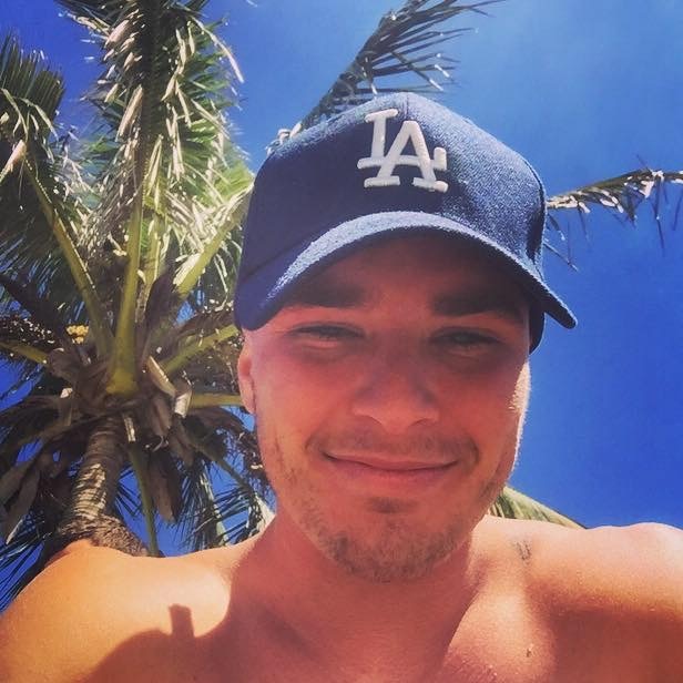 A smiling Luke Hansen wearing a baseball cap with a palm tree behind him.