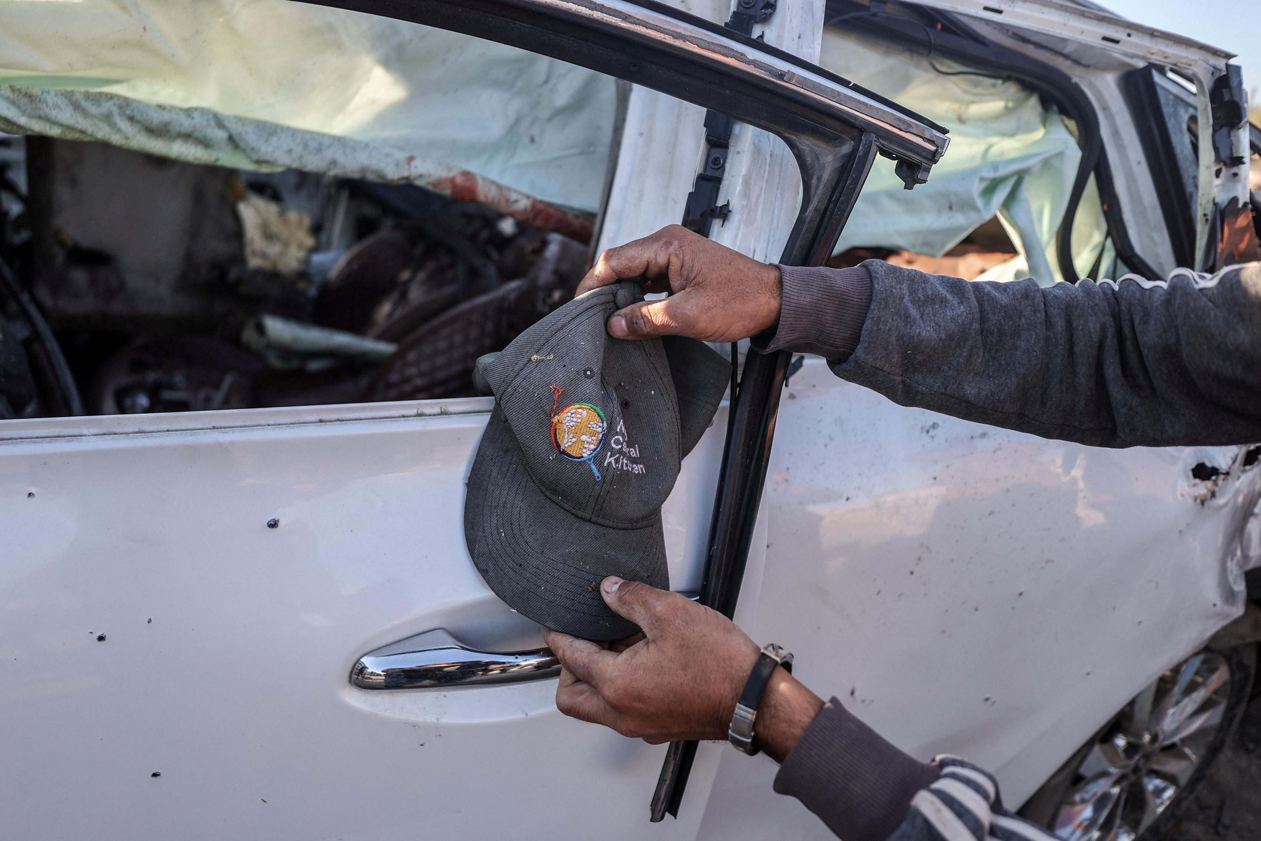 A photo of a man holding up a World Central Kitchen cap in front of a white car damaged by an air strike.