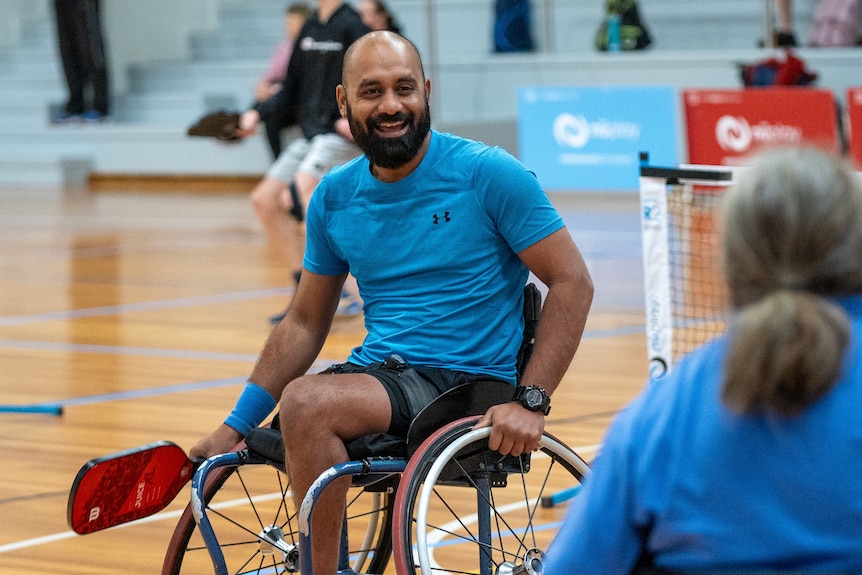 A man in a blue shirt in a wheelchair smiles on the pickleball court holding a paddle.