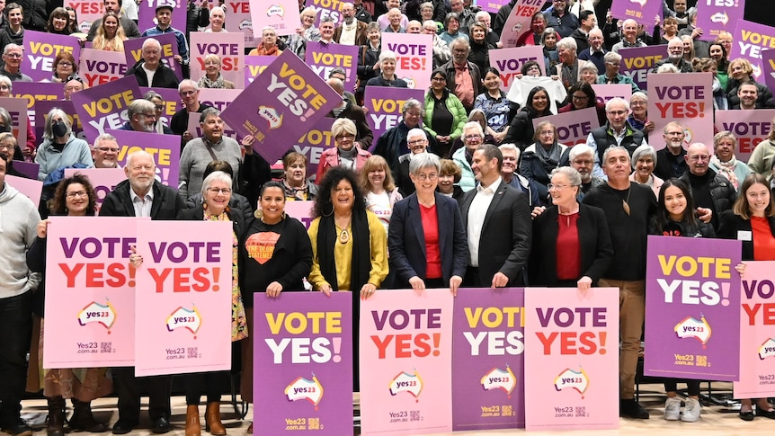 A large group of people in a school auditorium hold Vote Yes 23 signs
