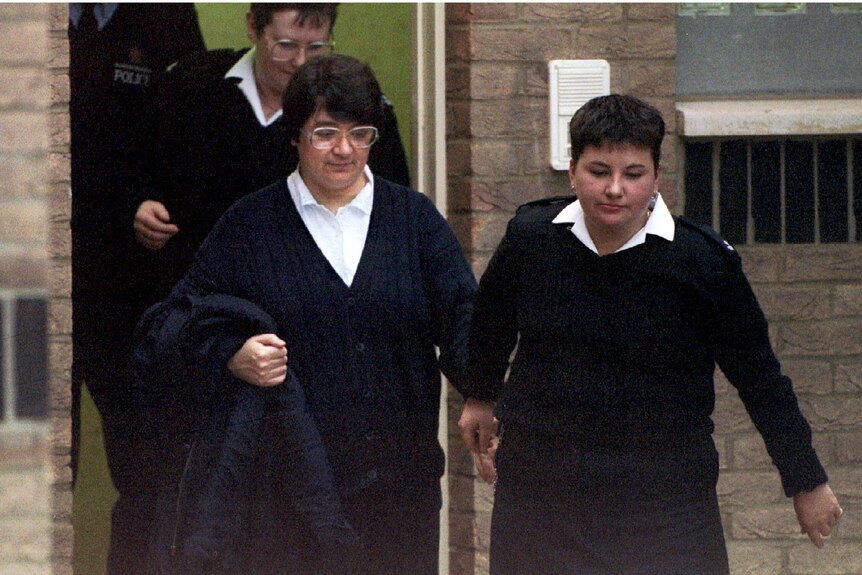 in a white blouse and cardigan is led out of a brick building by police