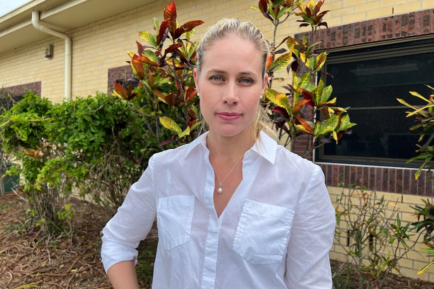 A blonde woman stands in front of a colourful bush, staring directy at the camera