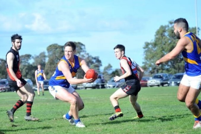 An action photo of an amateur Australian rules football match. A young man holds the ball, while surrounded by other players.