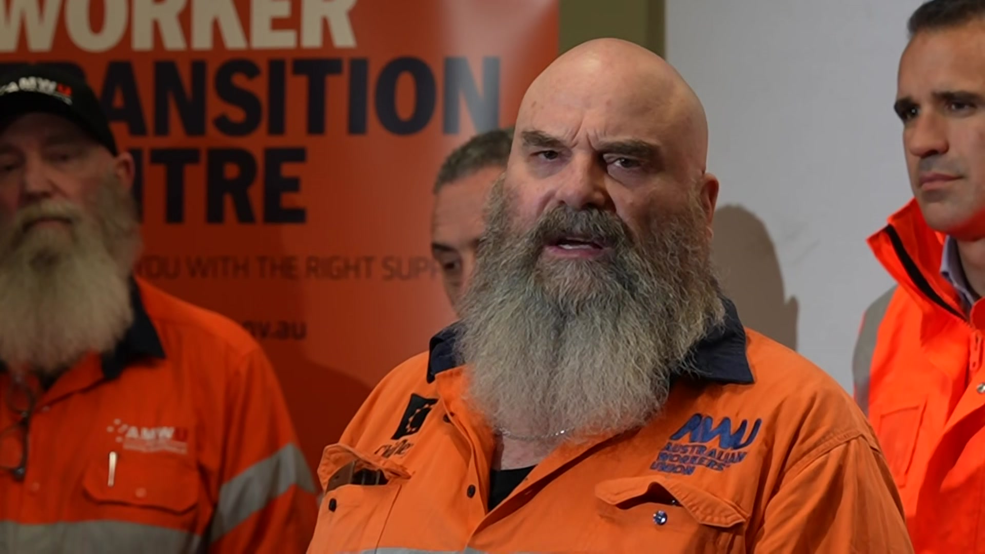 A man with a beard is standing before a row of people wearing orange work tops.