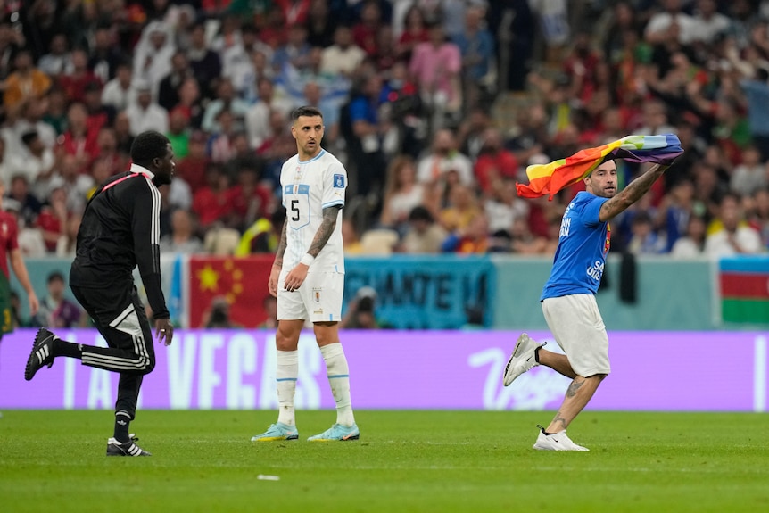 A referee chases a man carrying a rainbow flag while a soccer player watches