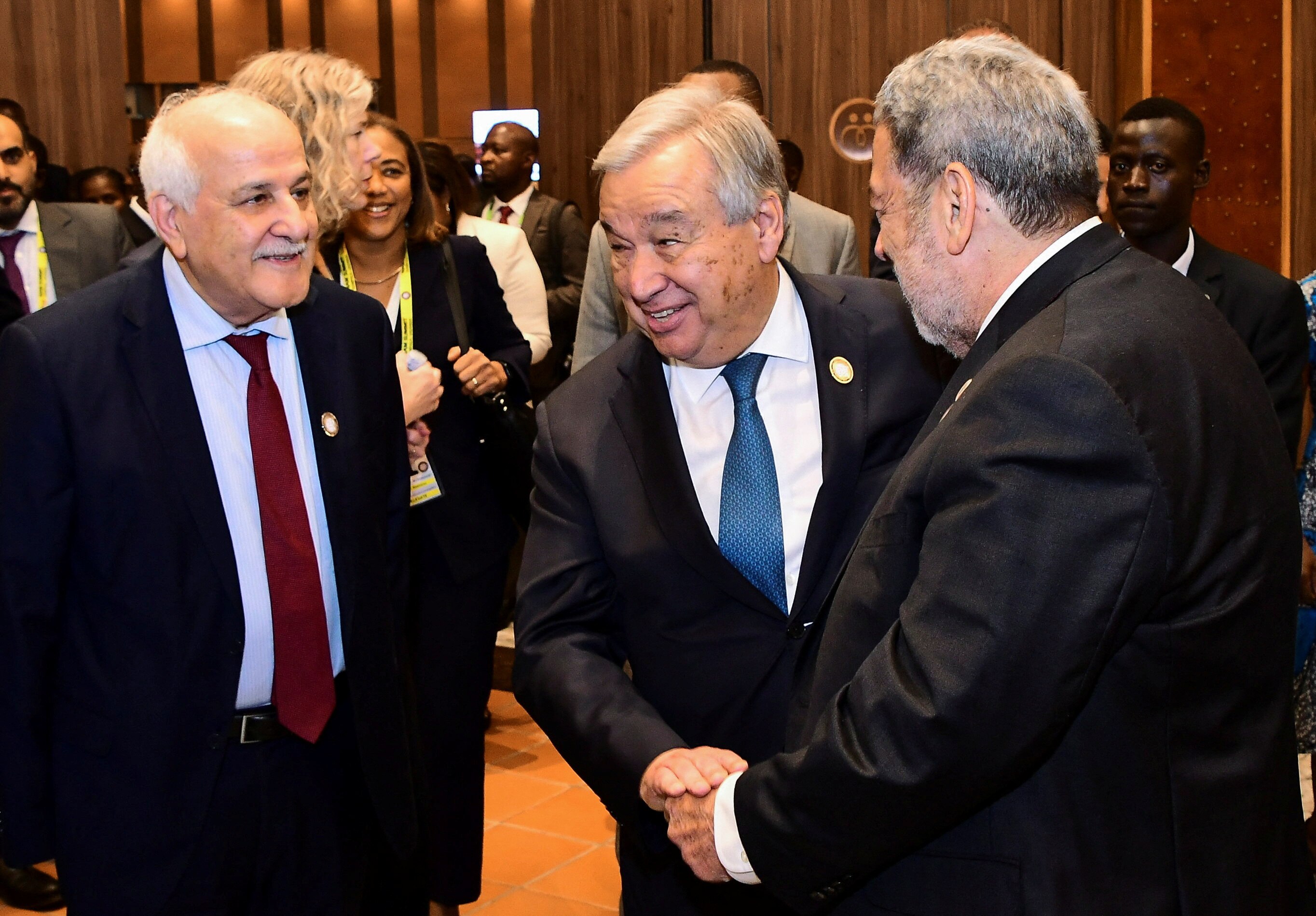 Three older men in suits stand smiling and shaking hands in a crowded, wood-panelled room.
